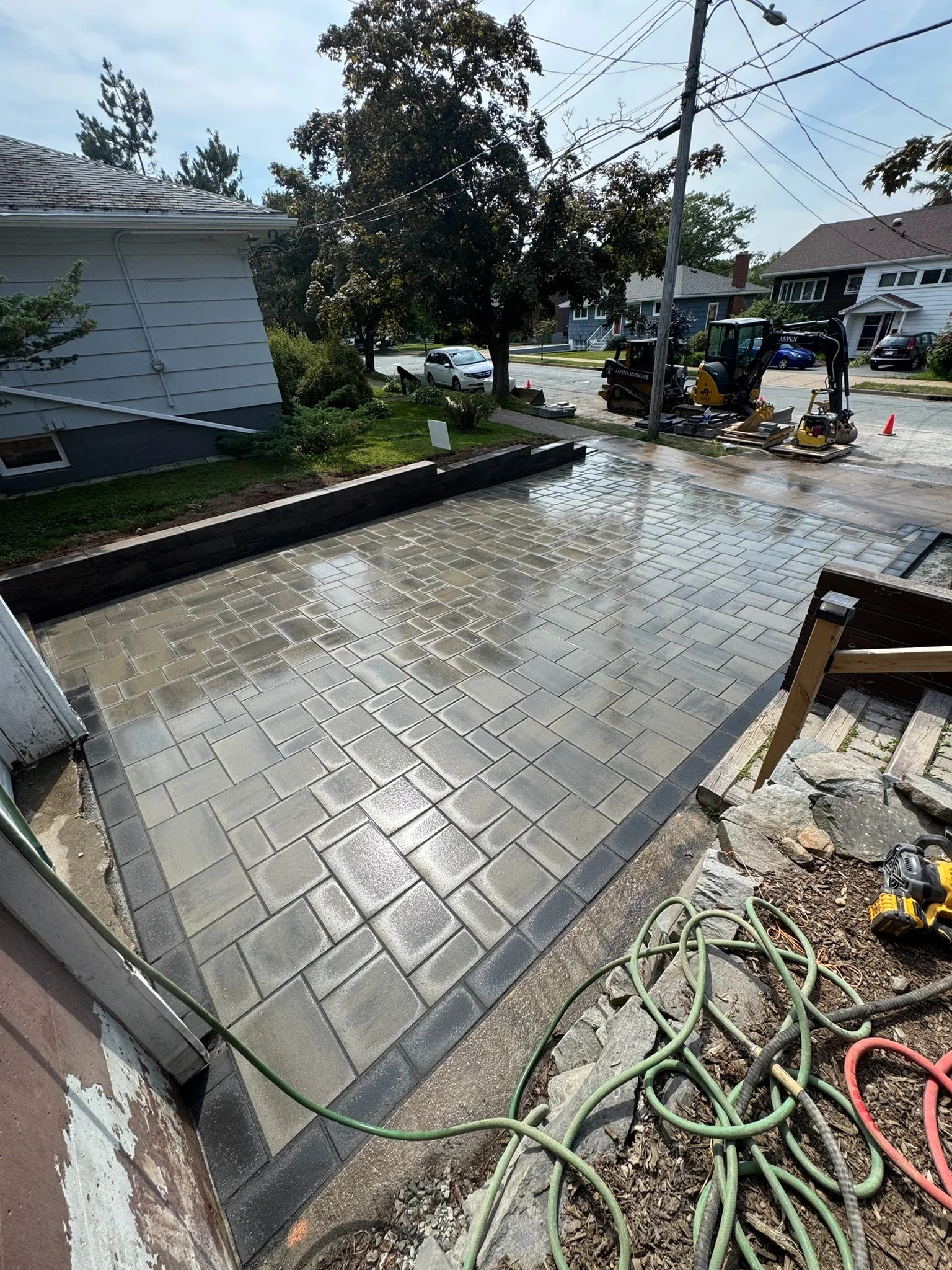 Newly paved concrete patio with stone border, construction tools and hoses, residential neighborhood street in background.