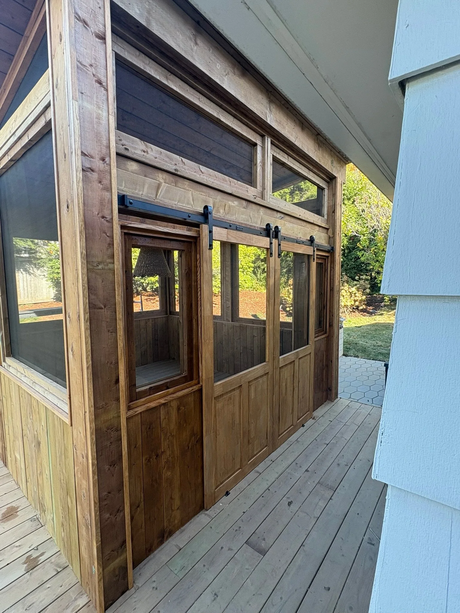Wooden screened porch with black barn door hardware, attached to a white house with a wooden deck and hexagonal tile pathway, surrounded by greenery.