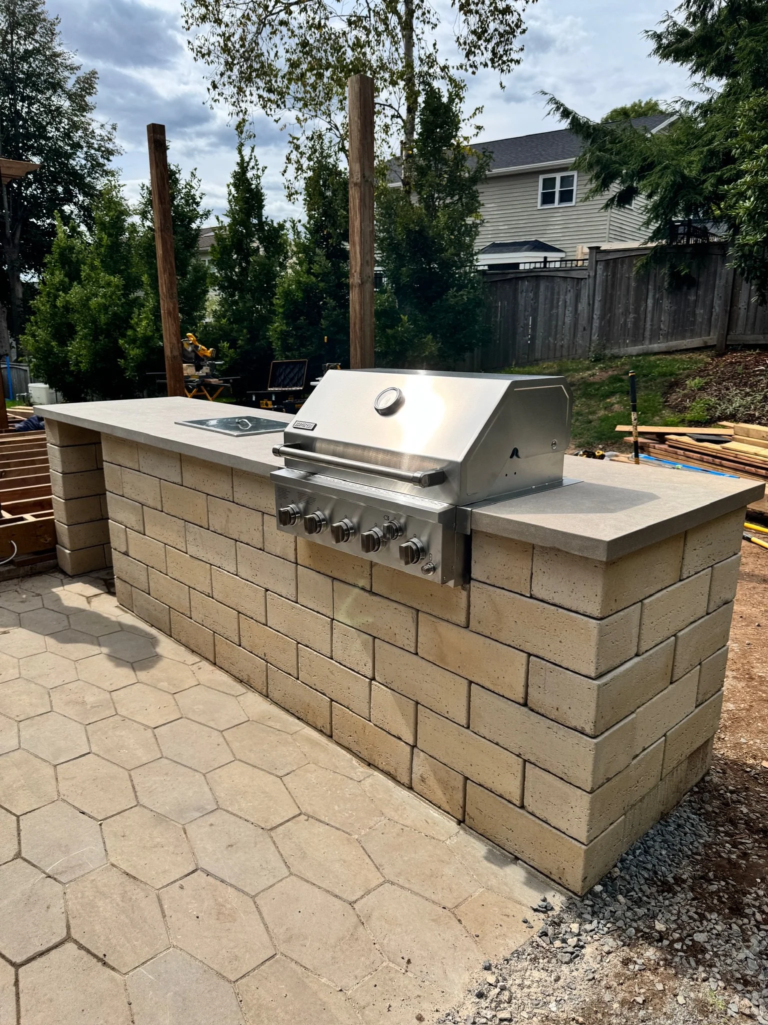 Outdoor kitchen featuring a built-in stainless steel gas grill on a brick counter with a concrete top, set in a backyard with trees and a wooden fence.