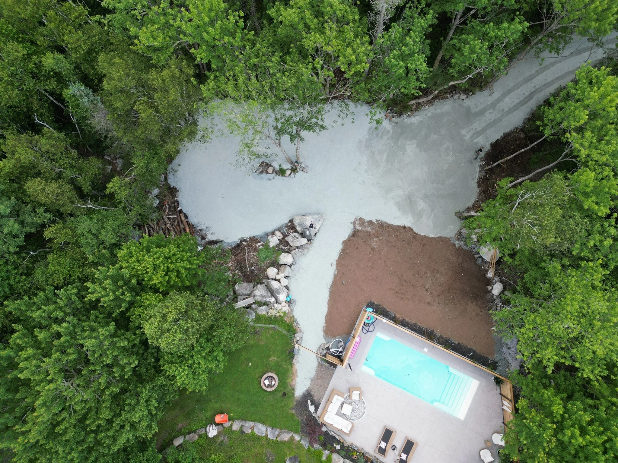 An aerial view of a backyard with a pool, a grassy area, a fire pit, walkways, and a wooded area surrounding the yard.