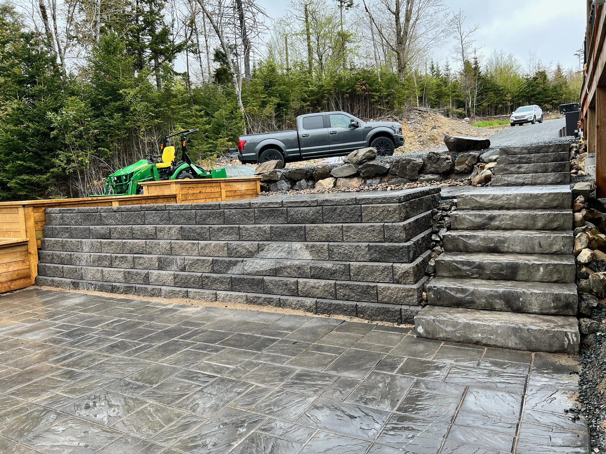 Outdoor patio with gray tiled floor, stone retaining wall, and stone staircase leading up to a gravel and dirt area with parked trucks, cars, and outdoor equipment, with trees in the background.