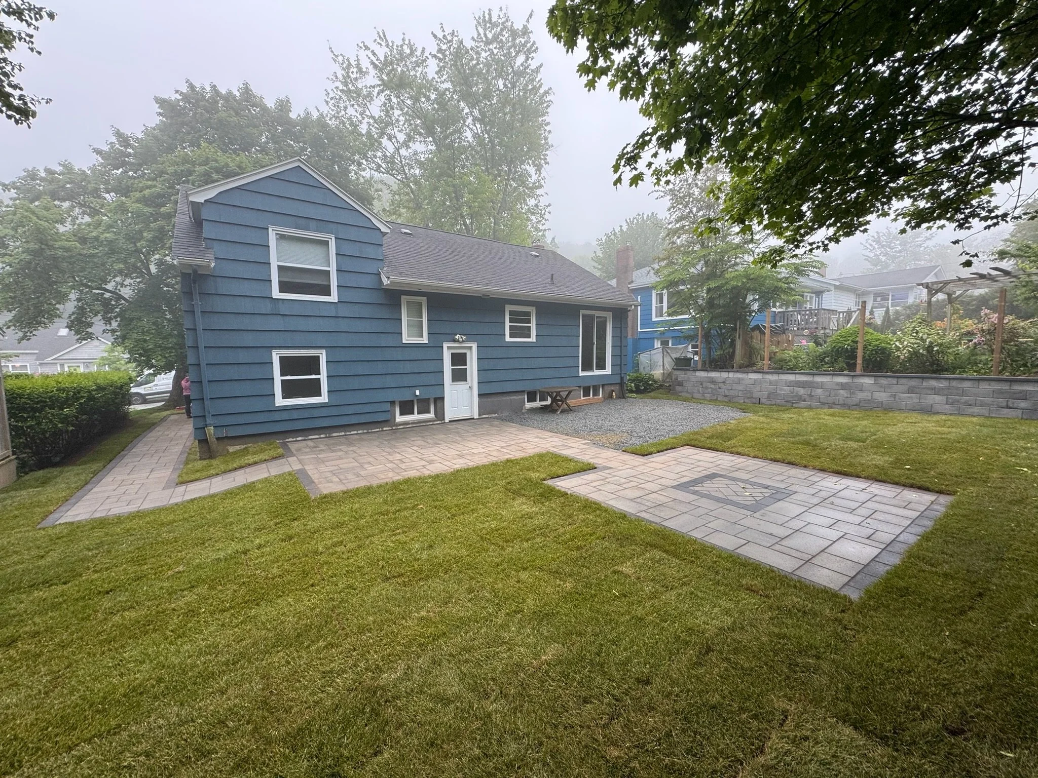 A blue two-story house with white window frames, a gravel section, and a paved patio area with a fire pit, surrounded by a well-maintained lawn and trees, on a foggy day.