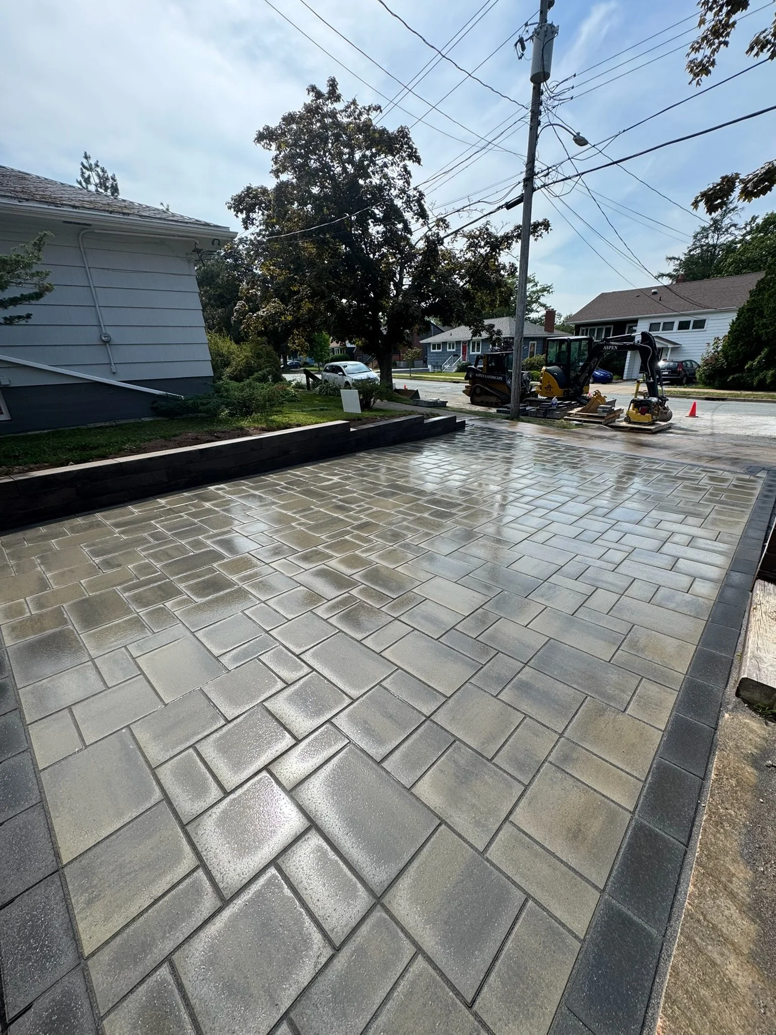 Recently paved concrete driveway with wet, shiny surface, bordered by wooden retaining wall and garden bed, during daytime with overcast sky.
