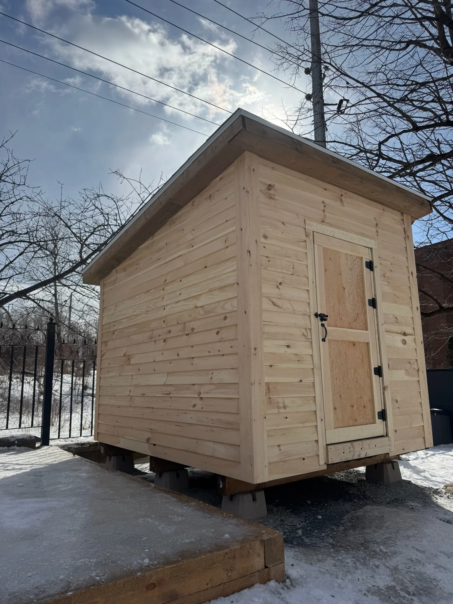 A small wooden shed with a sloped roof, built on concrete blocks, surrounded by snow, with a metal fence and leafless trees in the background.