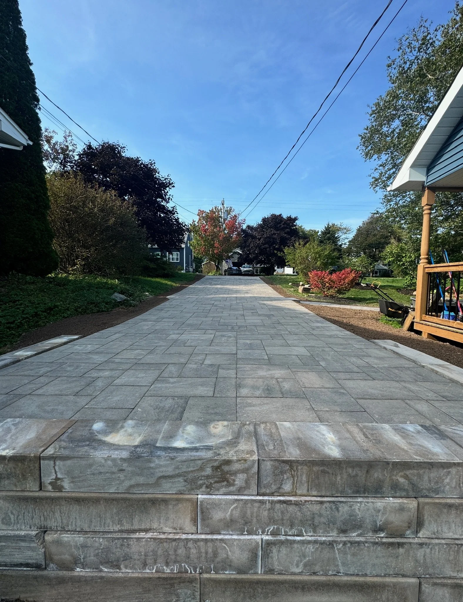 Newly paved driveway leading to a residential street under a blue sky, with trees and houses on either side.