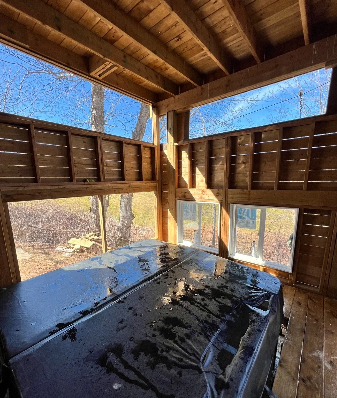 Interior view of a wooden structure under construction with view of blue sky and leafless trees outside, and a black object covered with plastic in the foreground.