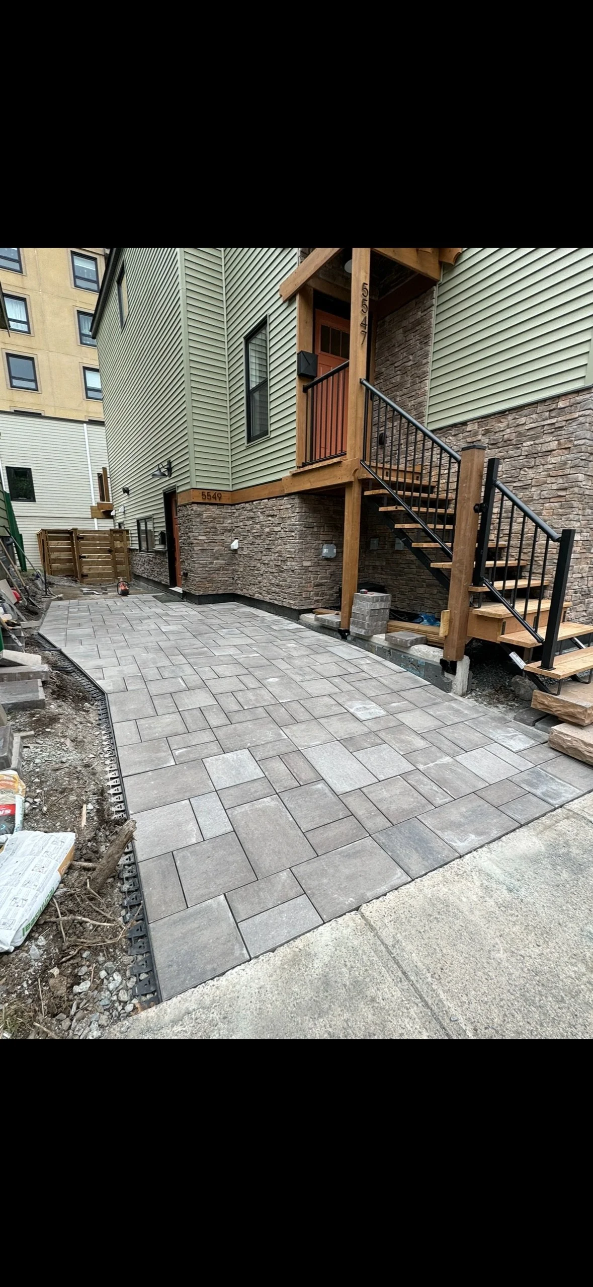 Newly installed stone paver patio in front of house with exterior stairs and green siding, construction materials and tools nearby.