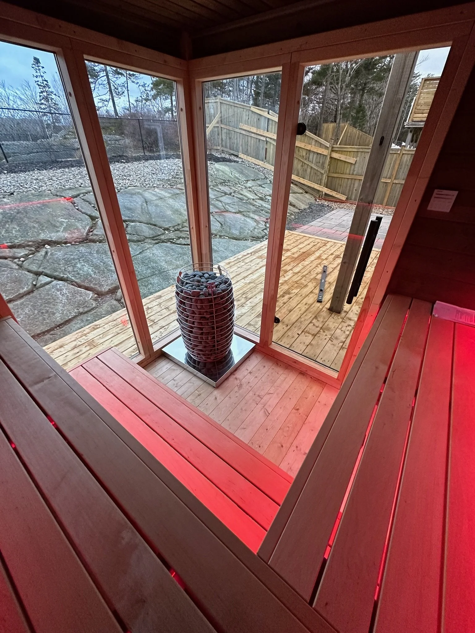 Inside a wooden sauna with a view of a small outdoor deck, glass doors, and rocky landscape outside, with a basket of dark stones inside.