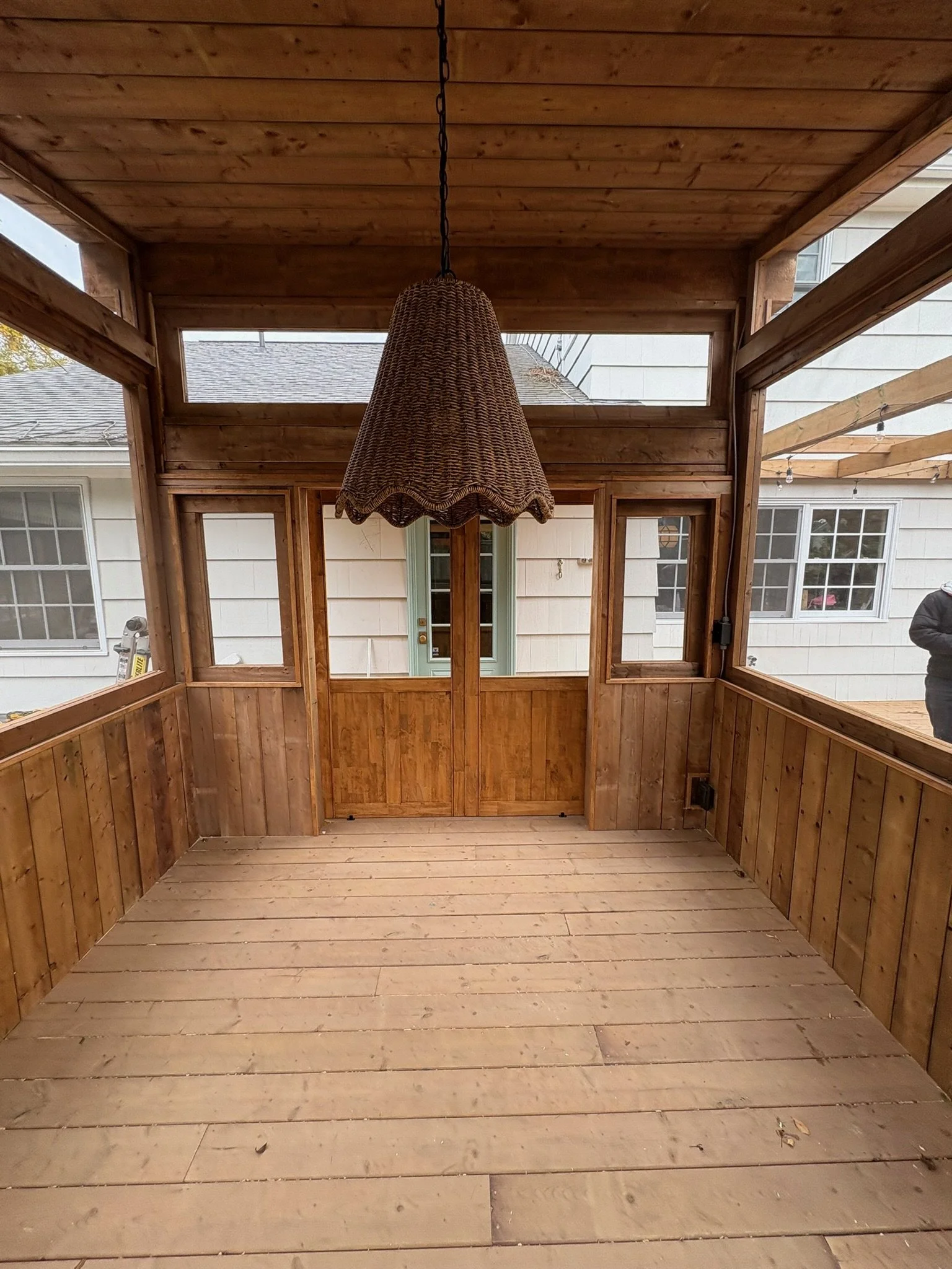 View of a wooden porch or sunroom with unfinished wooden flooring, wooden walls, a woven hanging lamp, and windows showing the outside of a house with white siding.