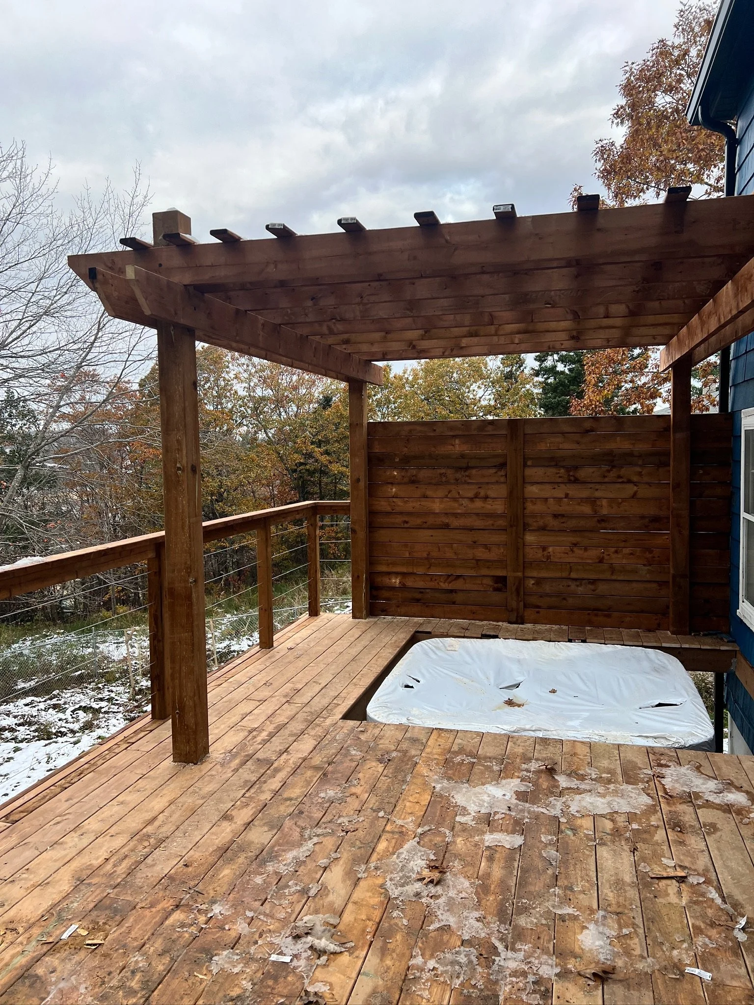 Wooden deck with a hot tub covered by a white plastic cover, surrounded by a weathered railing and a privacy fence, with trees and cloudy sky in the background.
