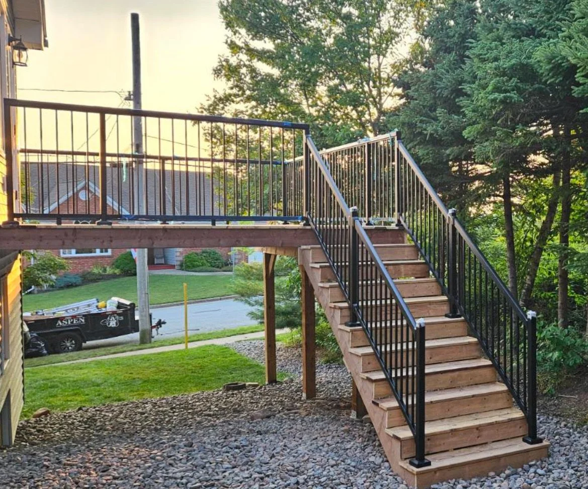 Wooden outdoor deck with metal railings and stairs, surrounded by trees and grass, in a residential neighborhood during sunset.