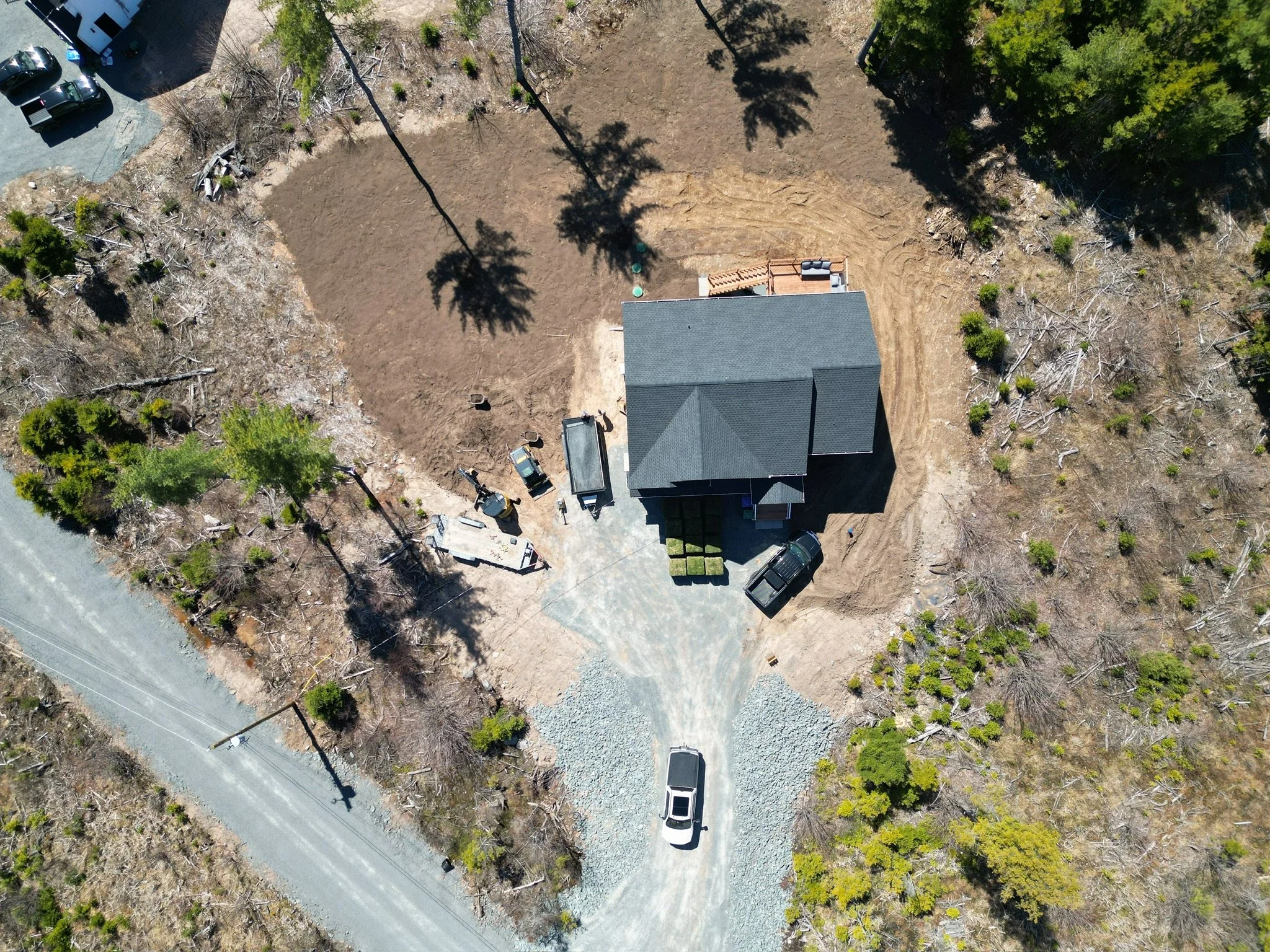 An aerial view of a house under construction with a gray roof, surrounded by trees, a gravel driveway with parked trucks and construction equipment, and a partially cleared yard.
