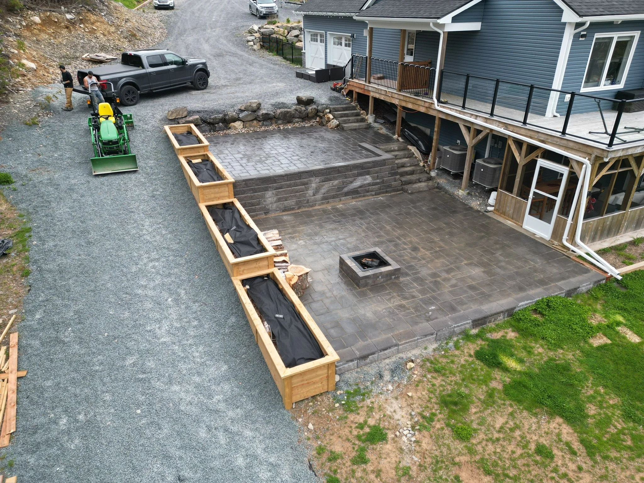 An overview of a house with a newly constructed outdoor patio, including stairs, a fire pit, and surrounding planters. Two men are near a black pickup truck, with gardening or construction equipment, on a gravel driveway.