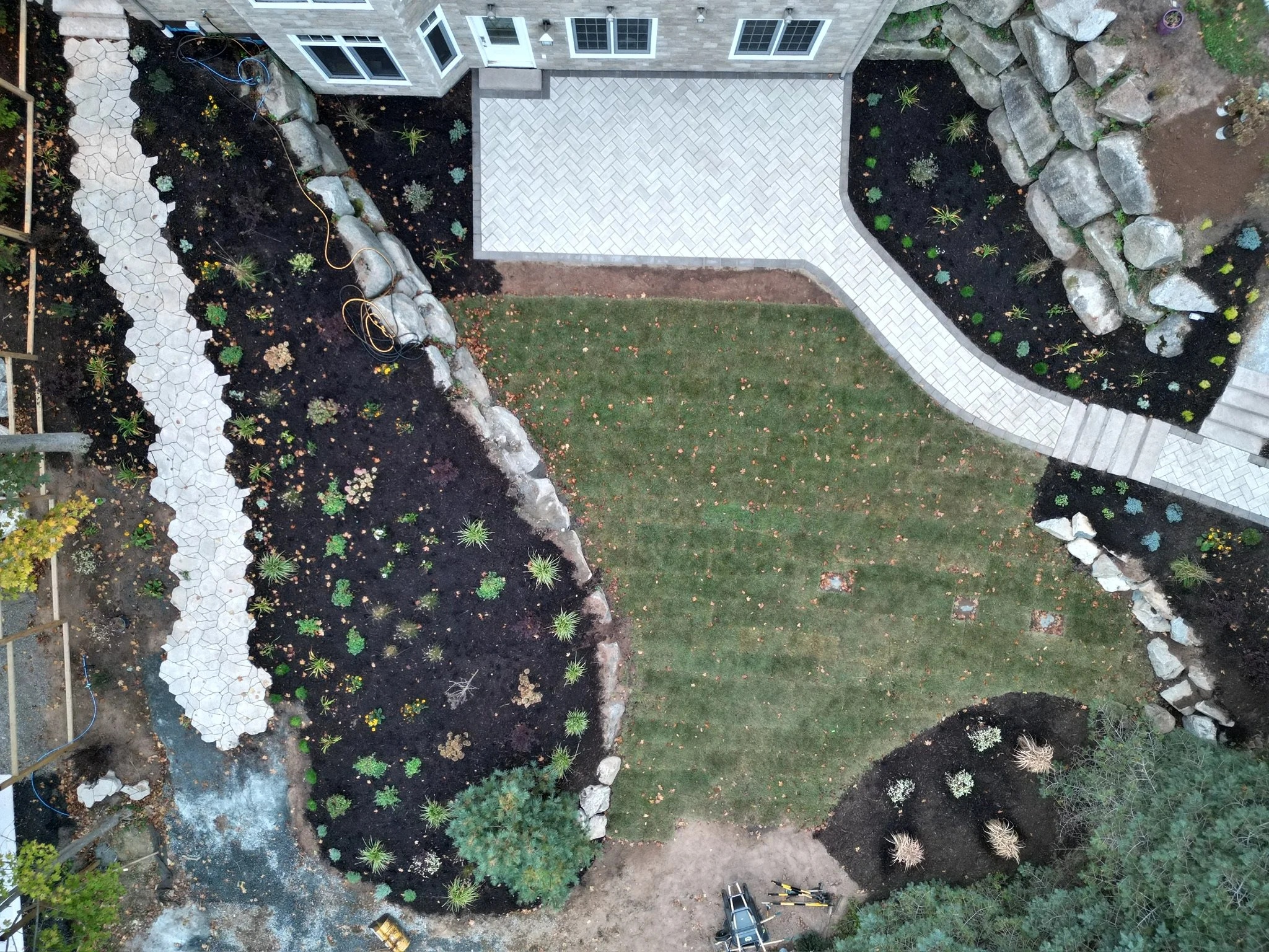 An aerial view of a landscaped backyard with a lawn, paved pathways, garden beds with rocks and plants, and a house with a balcony.