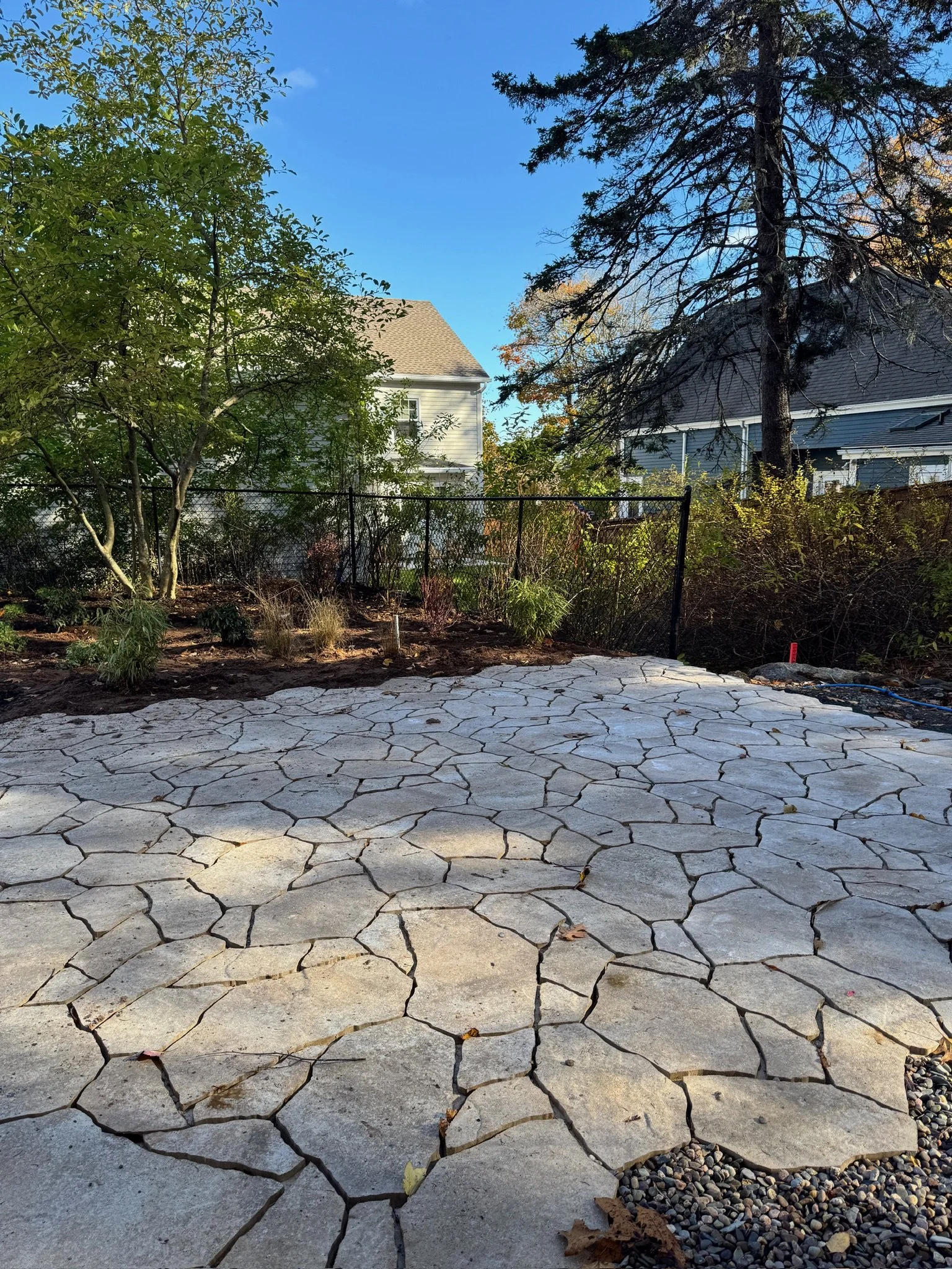 A backyard with a newly paved stone patio, surrounded by trees and a black metal fence, with houses visible in the background.