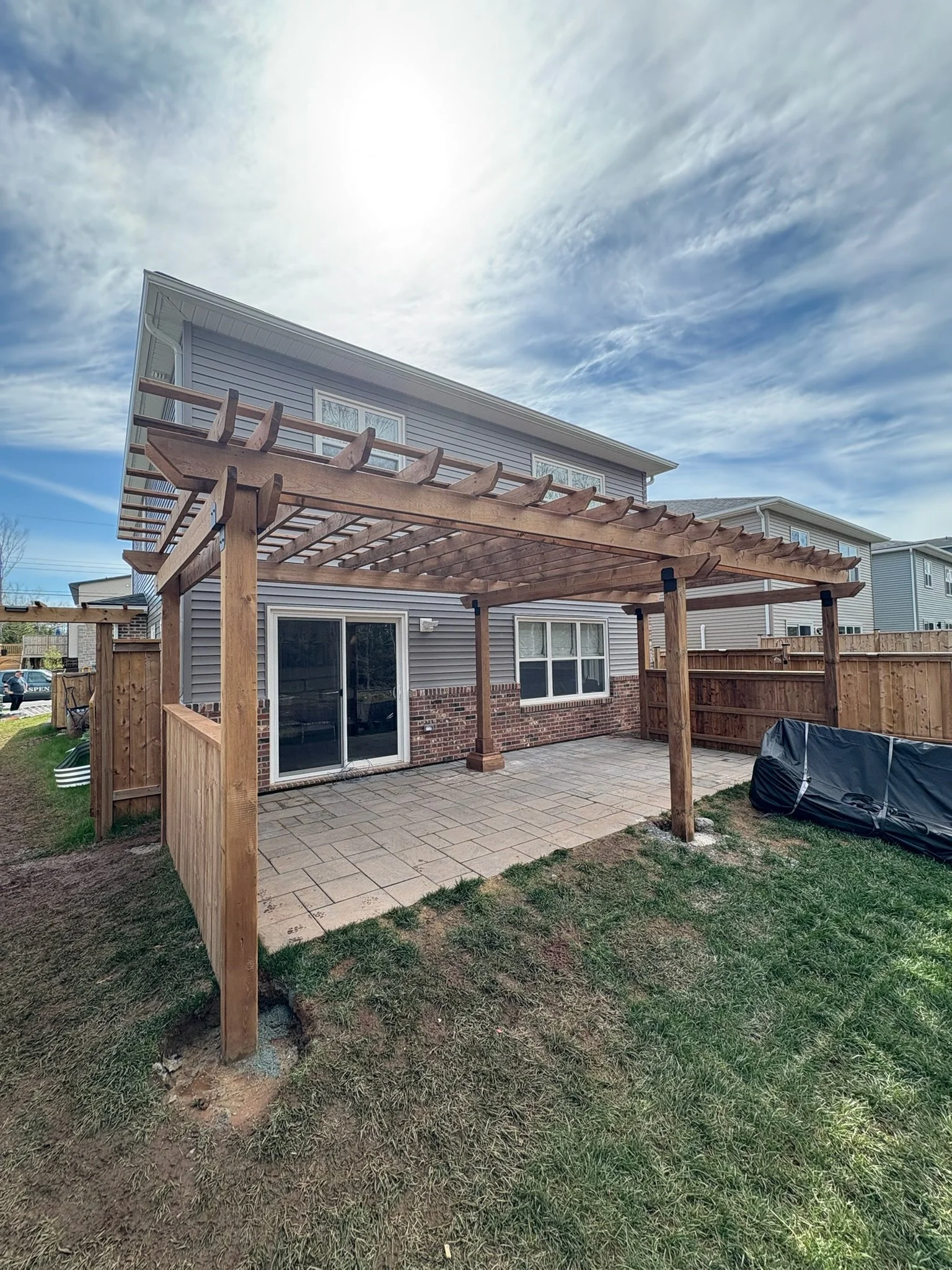 Backyard patio with a newly built wooden pergola and sliding glass door, lawn, and neighboring houses under a partly cloudy sky.