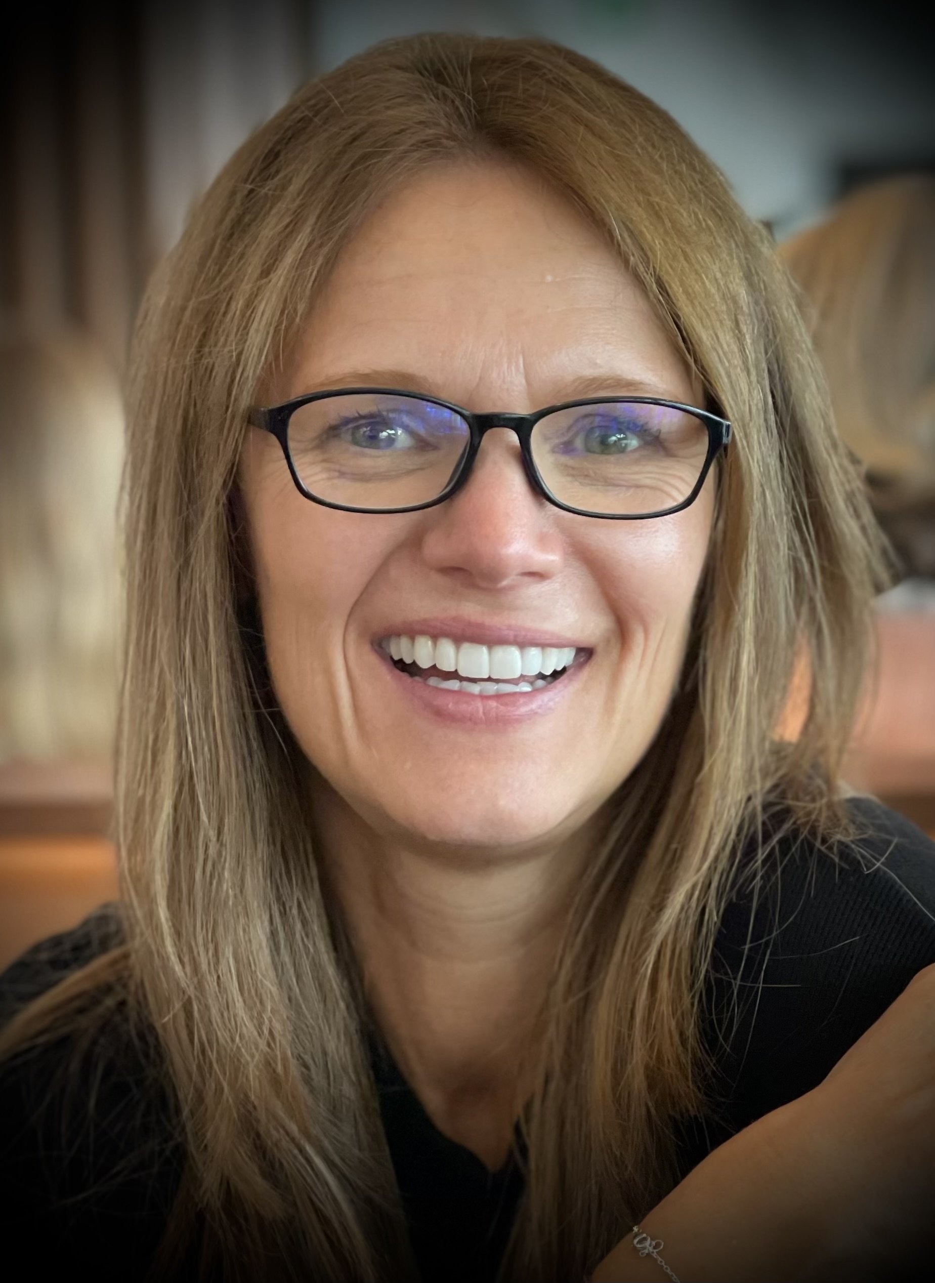 Close-up of a smiling woman with long, light brown hair, wearing glasses, in an indoor setting.