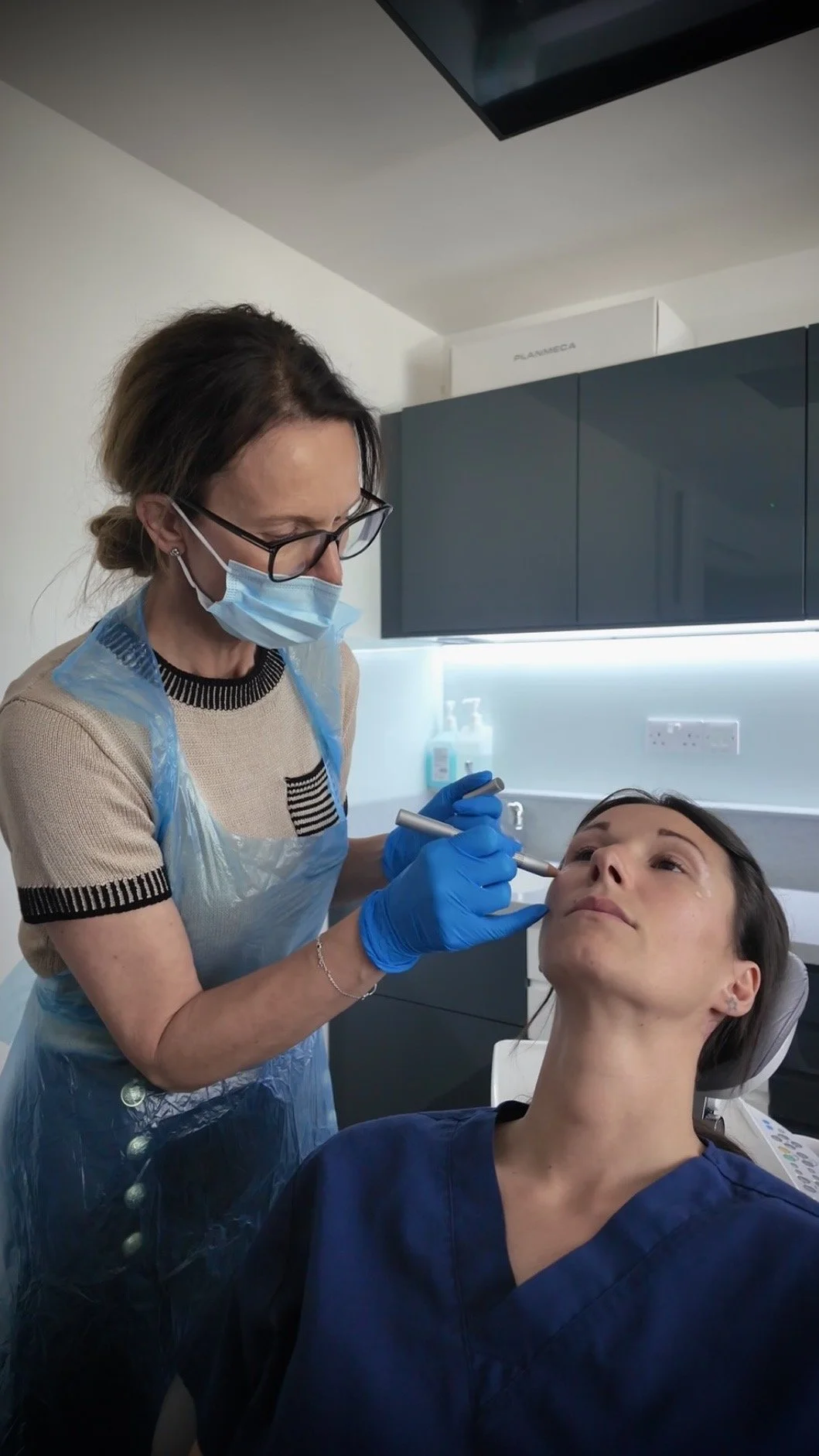 Dentist in blue gloves and face mask performing a procedure on a woman's face in a dental office.