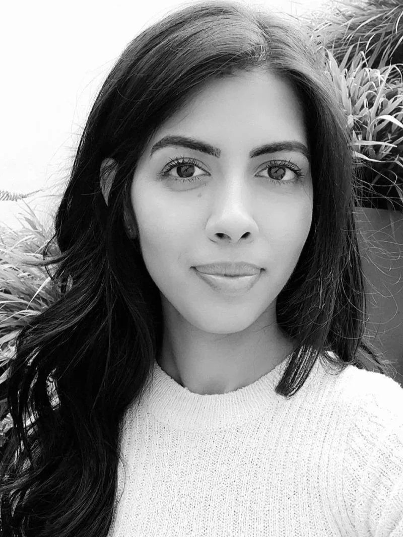 A black-and-white photo of a young woman with long dark hair, wearing a light-colored sweater, standing outdoors with plants in the background.