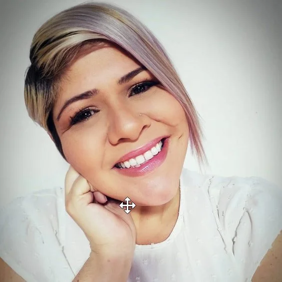 A woman with short, pastel-colored hair smiling with her hand resting on her cheek.