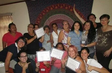 Group of women and girls indoors, some holding certificates, celebrating together, with a decorative tapestry in the background.
