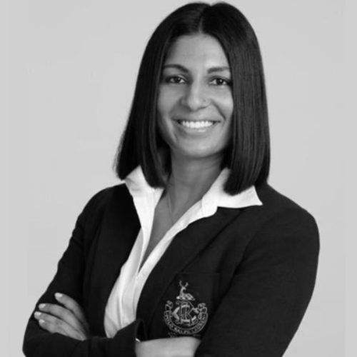 Professional woman with shoulder-length dark hair, smiling, wearing a blazer with an emblem and a white blouse in a studio portrait.