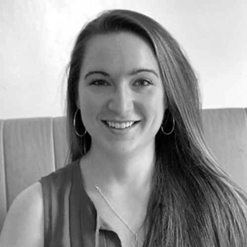 Black and white portrait of a smiling woman with long hair, wearing earrings and a sleeveless top, sitting in front of a padded headboard.