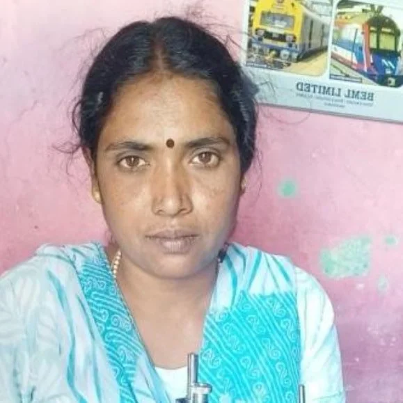 A woman with dark hair, a bindi on her forehead, wearing a white and blue traditional Indian dress, sitting in front of a pink wall with a poster of buses behind her.