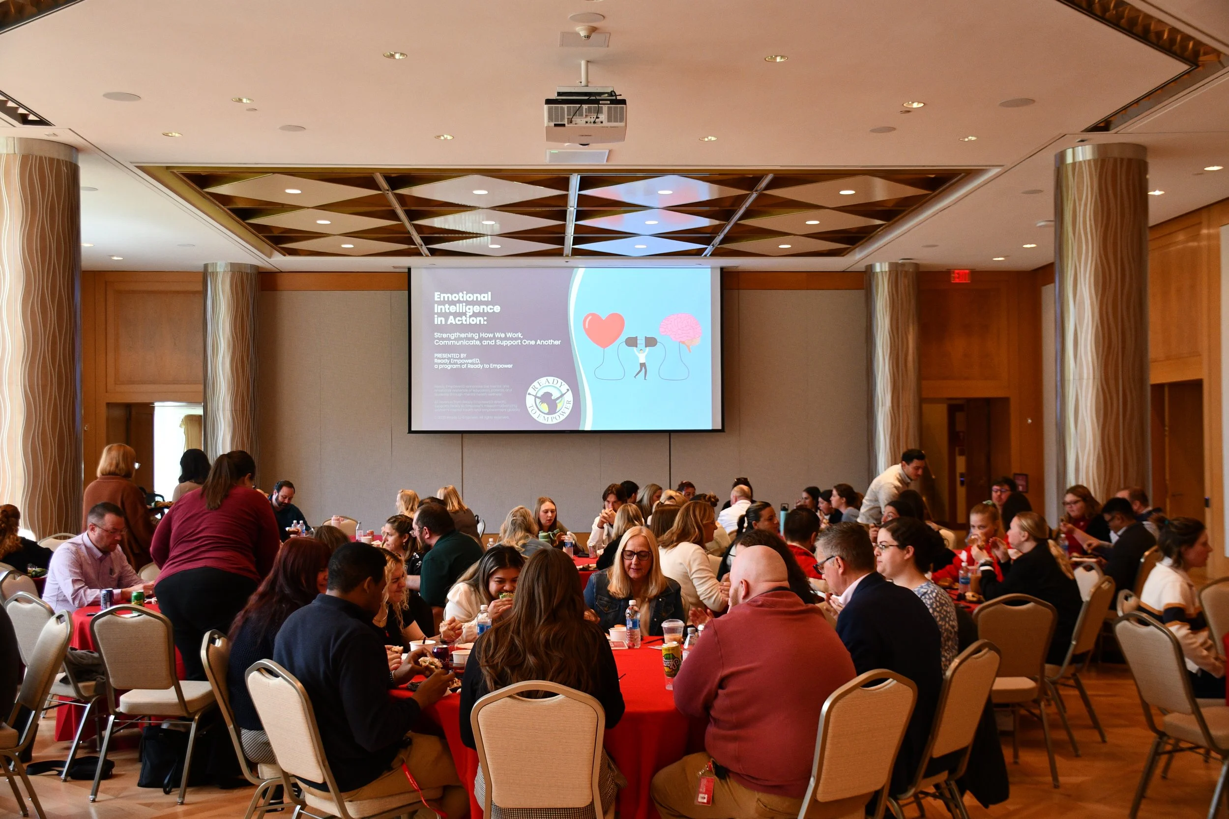 A large conference room filled with people sitting at round tables covered with red tablecloths, listening to a presentation on emotional intelligence displayed on a large screen at the front of the room.