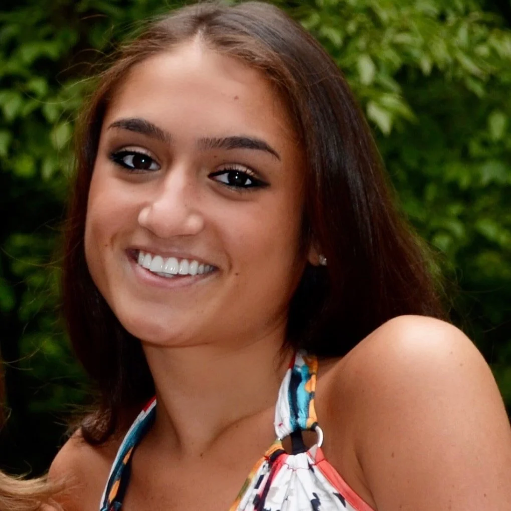 A young woman with brown hair, smiling, wearing a colorful sleeveless top, with a blurred green leafy background.