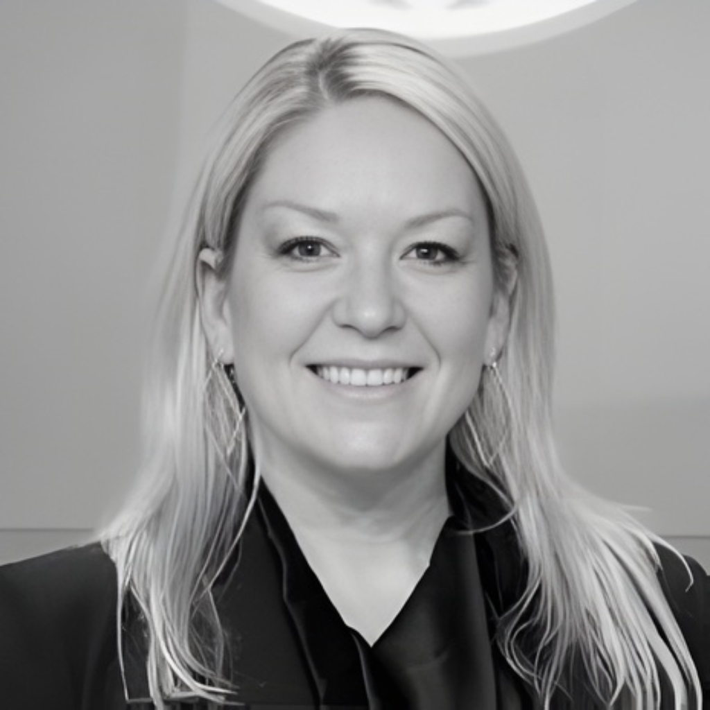 Black and white portrait of a smiling woman with long, light-colored hair, earrings, wearing a dark blazer and a top, against a plain background.