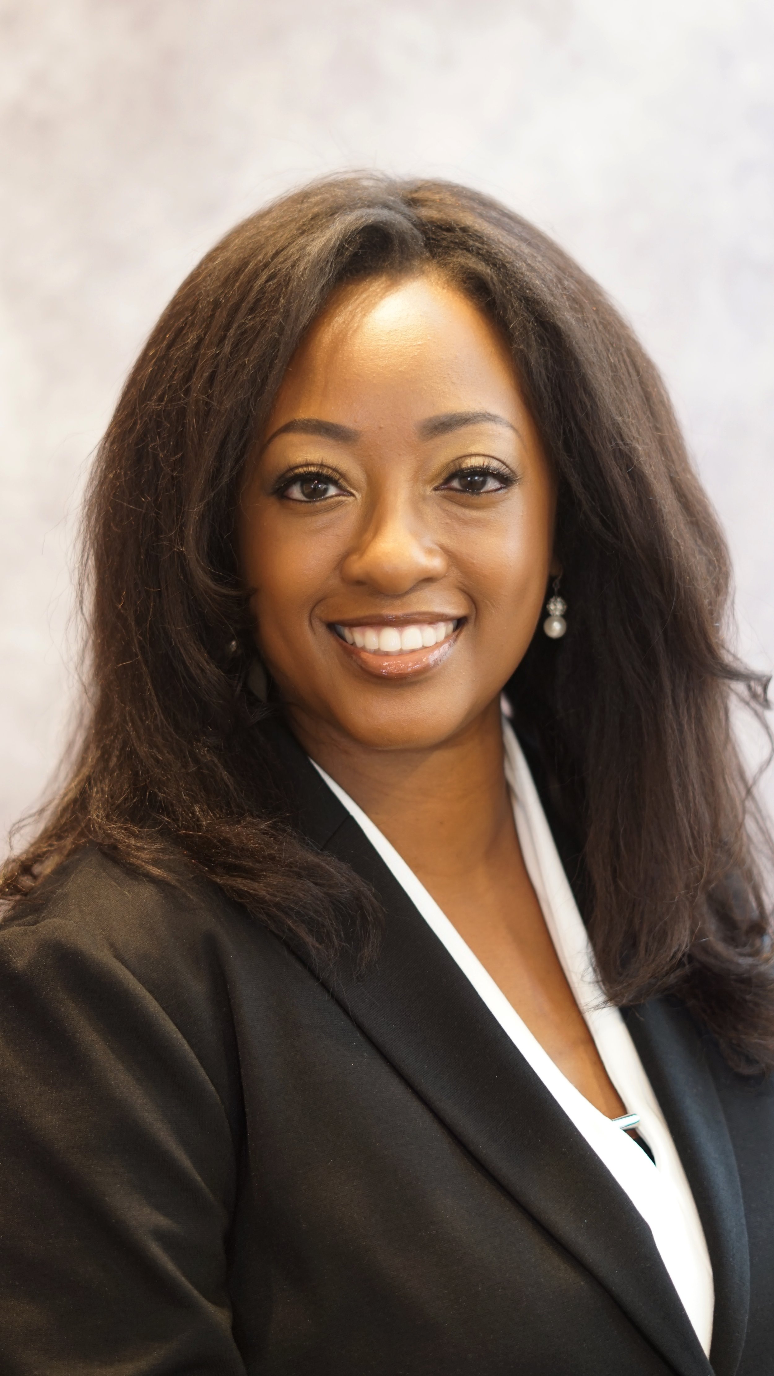 A professional woman with long dark hair, wearing a black blazer and white blouse, smiling against a neutral background.