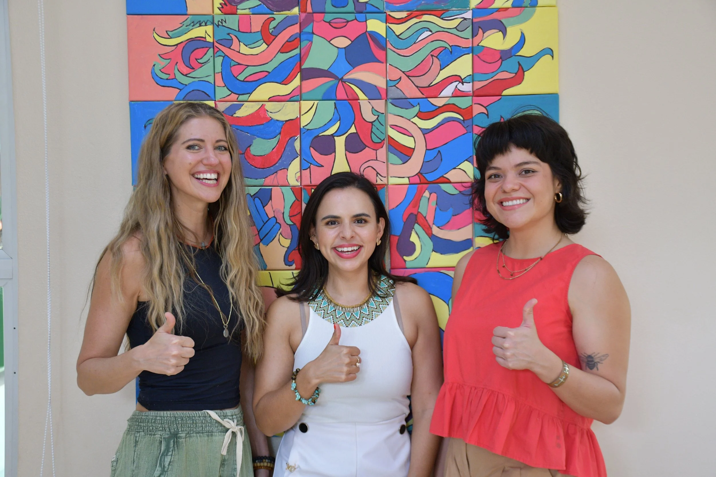 Three women standing in front of colorful abstract wall art, smiling and giving a thumbs-up.