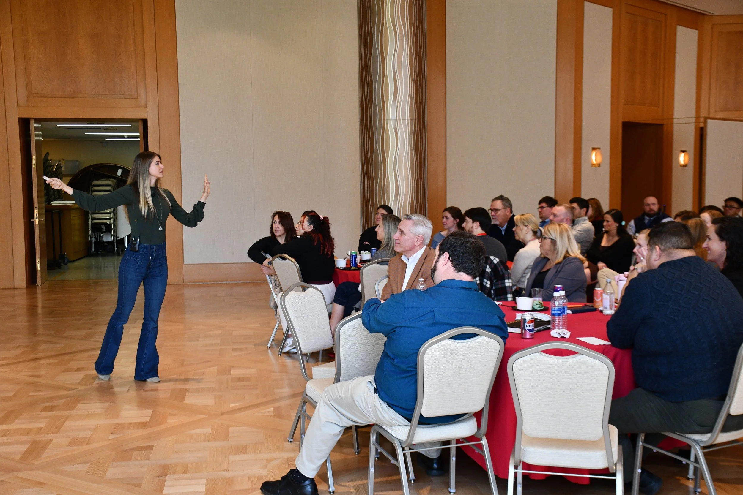 A woman standing in front of a seated audience presenting or speaking during a conference or seminar in a large room with wood-paneled walls and a polished wooden floor.