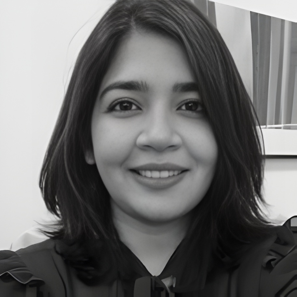 A smiling woman with shoulder-length dark hair, wearing a dark top, in front of a plain background with a partial view of a window or picture frame.