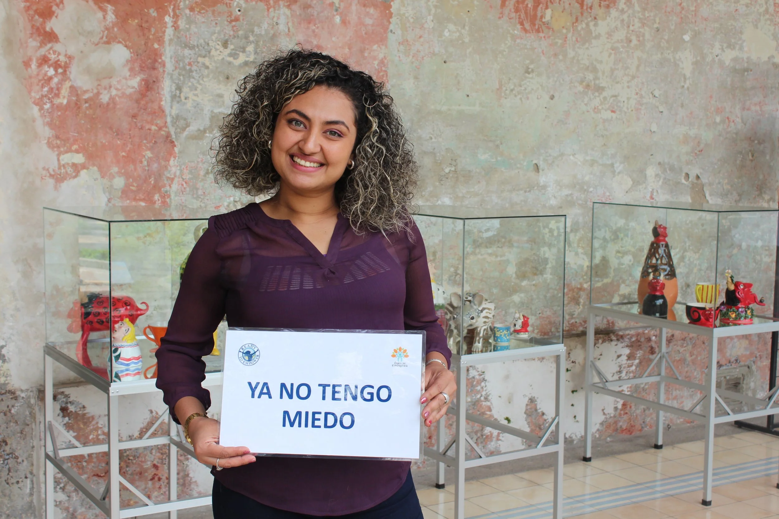 A woman with curly hair smiling and holding a sign that says 'YA NO TENGO MIEDO' in a room with colorful decorative objects displayed in glass cases.