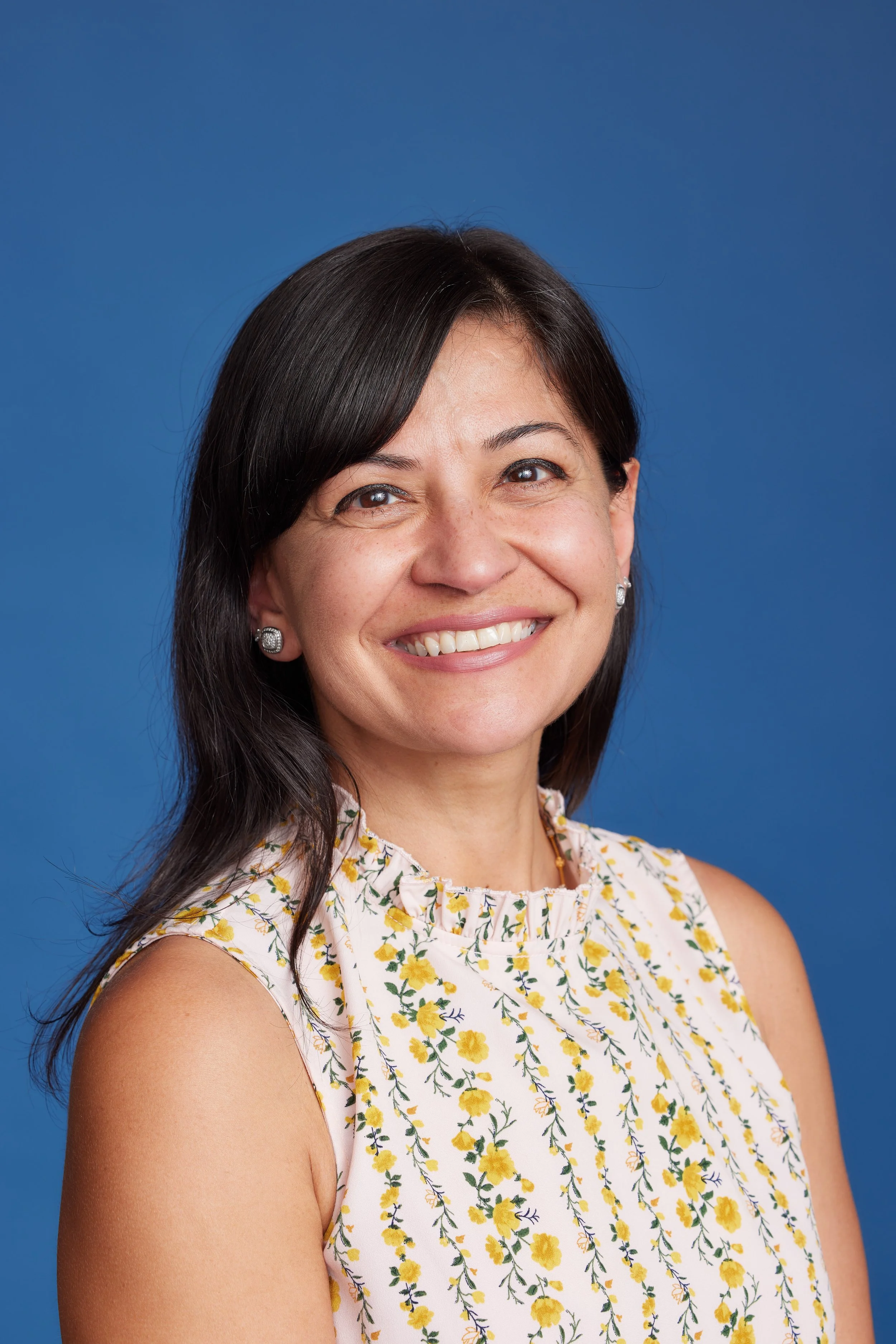 A woman smiling with dark hair, wearing a sleeveless floral top, earrings, and a necklace, against a blue background.
