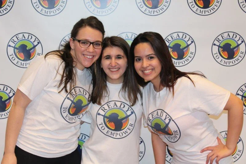 Three young women smiling and wearing white T-shirts that say "READY TO EMPOWER" with a logo, standing in front of a backdrop featuring the same logo.