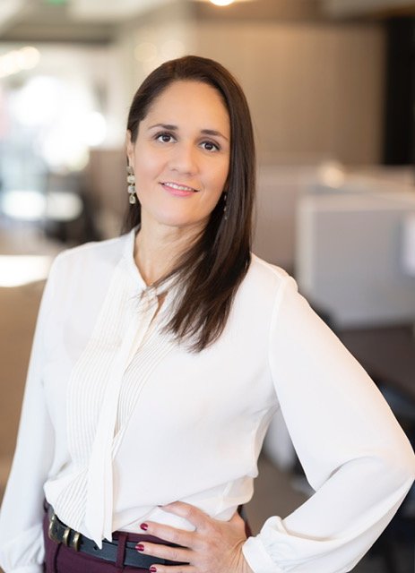 A woman with long brown hair, wearing a white blouse and dark pants, standing indoors with a smile.