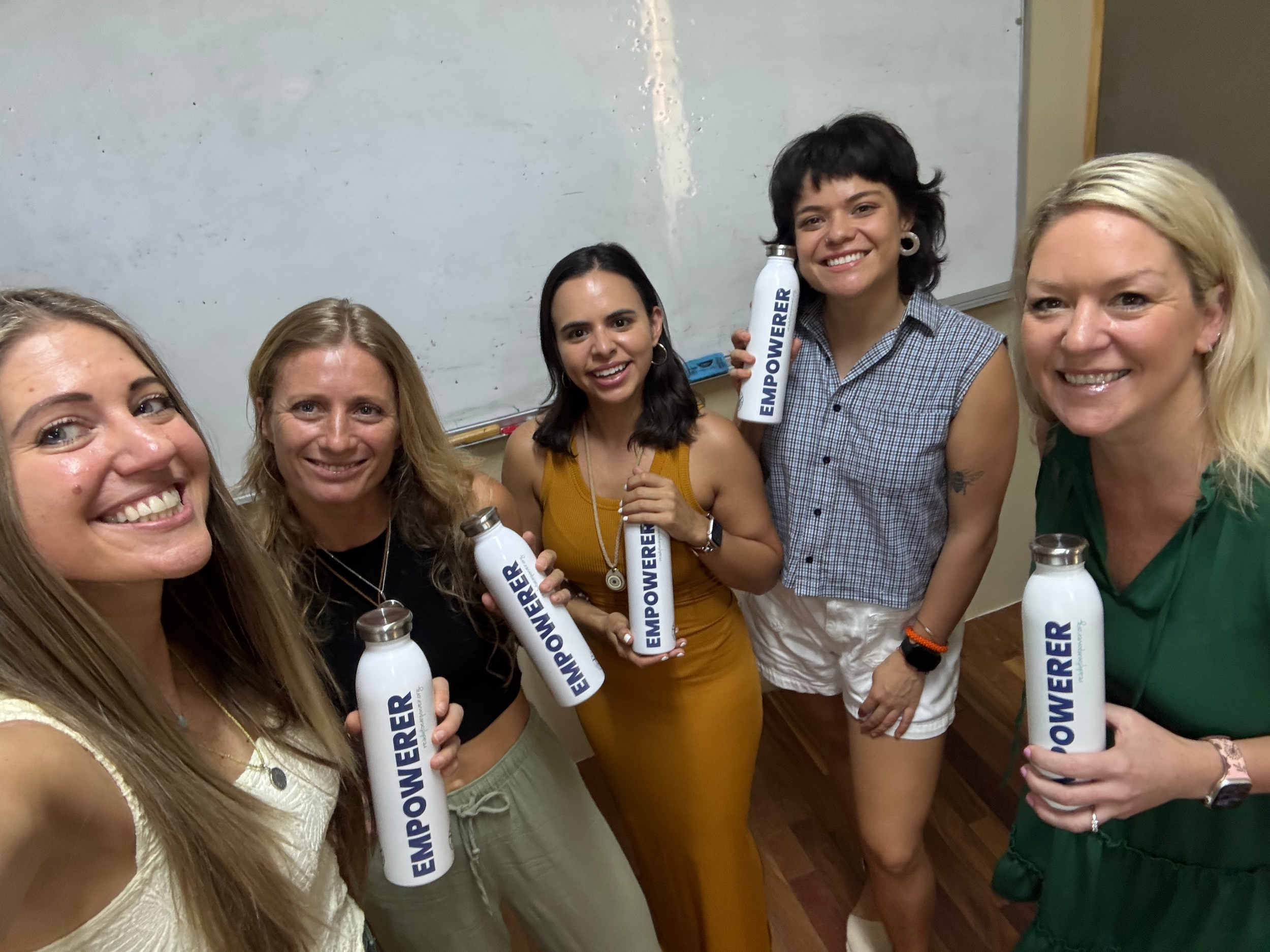 Five women smiling and holding white water bottles with 'Empower' written on them in a classroom with a whiteboard in the background.