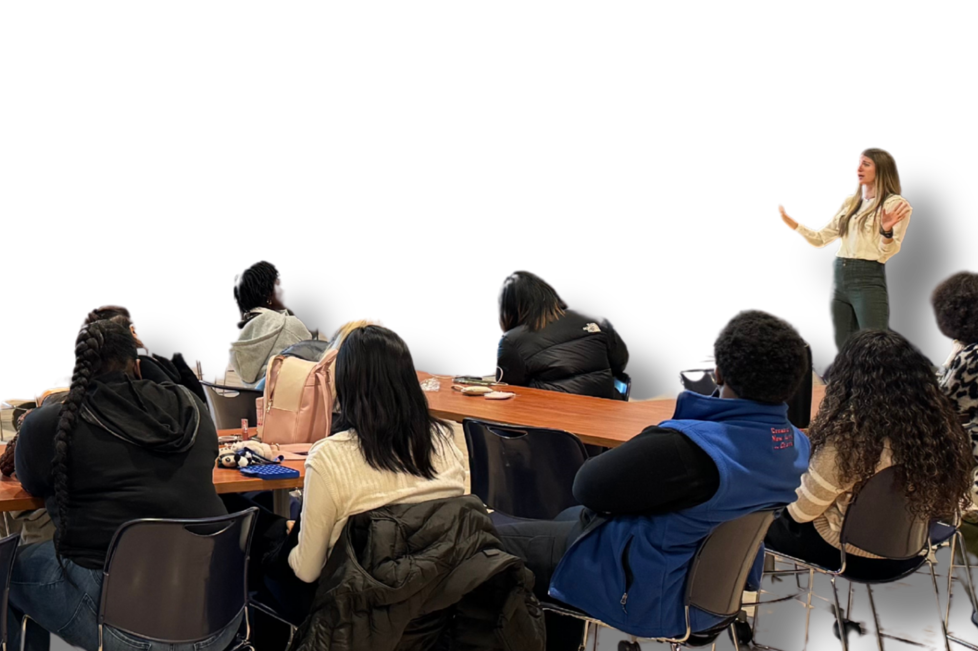 A classroom scene with a young woman standing and speaking to a diverse group of students seated at tables, some taking notes and others listening attentively.