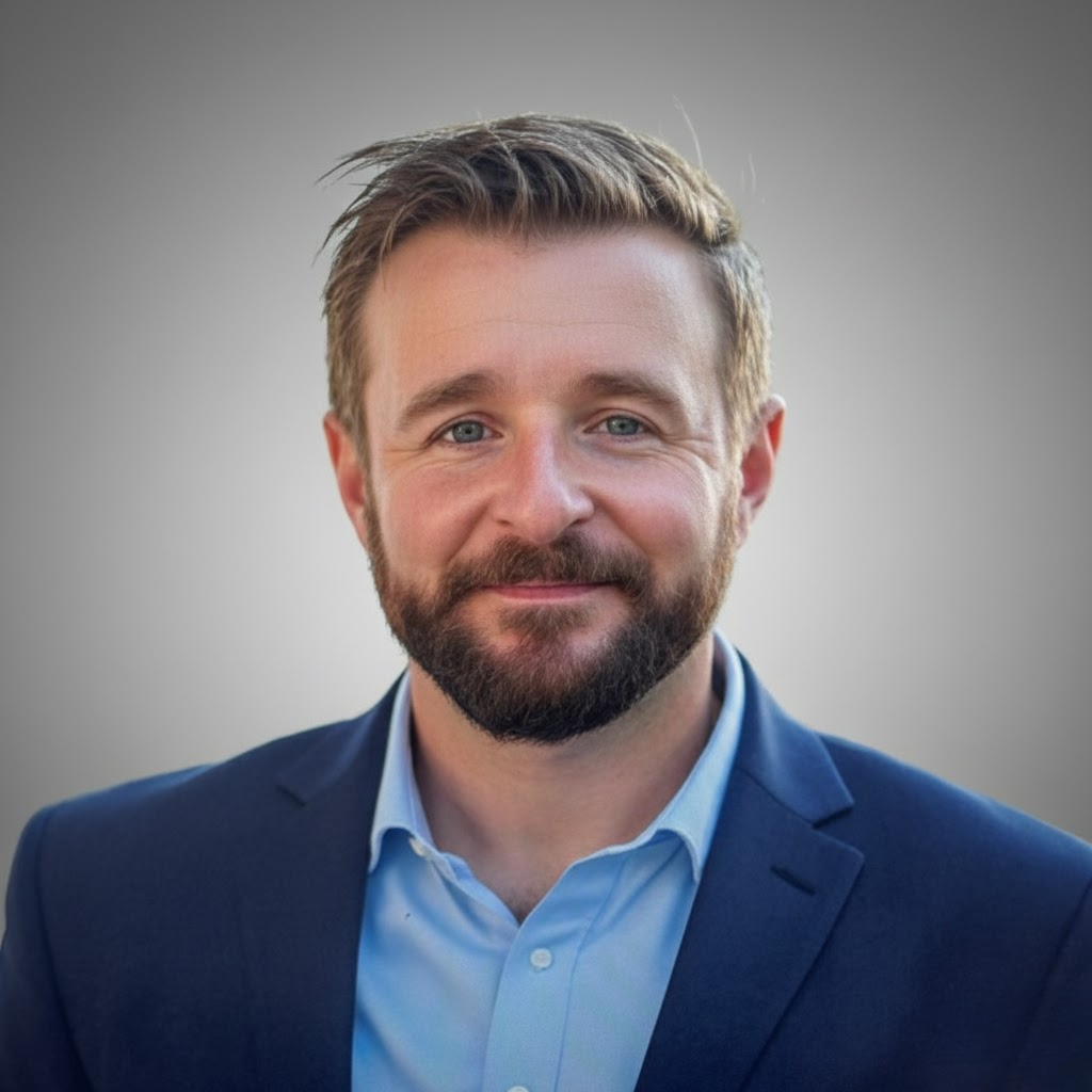 A man with light brown hair, a beard, and blue eyes, dressed in a navy blazer and light blue collared shirt, smiling at the camera with a blurred gray background.
