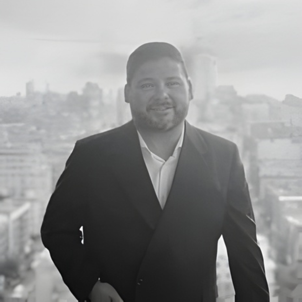 A man in a suit smiling and standing in front of a cityscape with tall buildings, possibly taken from a high vantage point or balcony.
