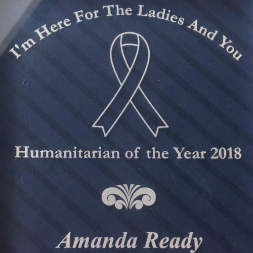 A poster with a blue background and diagonal stripes, featuring a white awareness ribbon and text that reads: "I'm Here For The Ladies And You," "Humanitarian of the Year 2018," and "Amanda Ready."