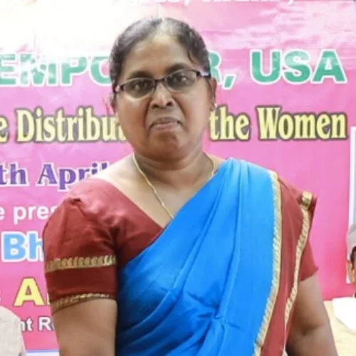 A woman wearing glasses and a traditional red and blue saree, standing in front of a pink banner with green and yellow text.