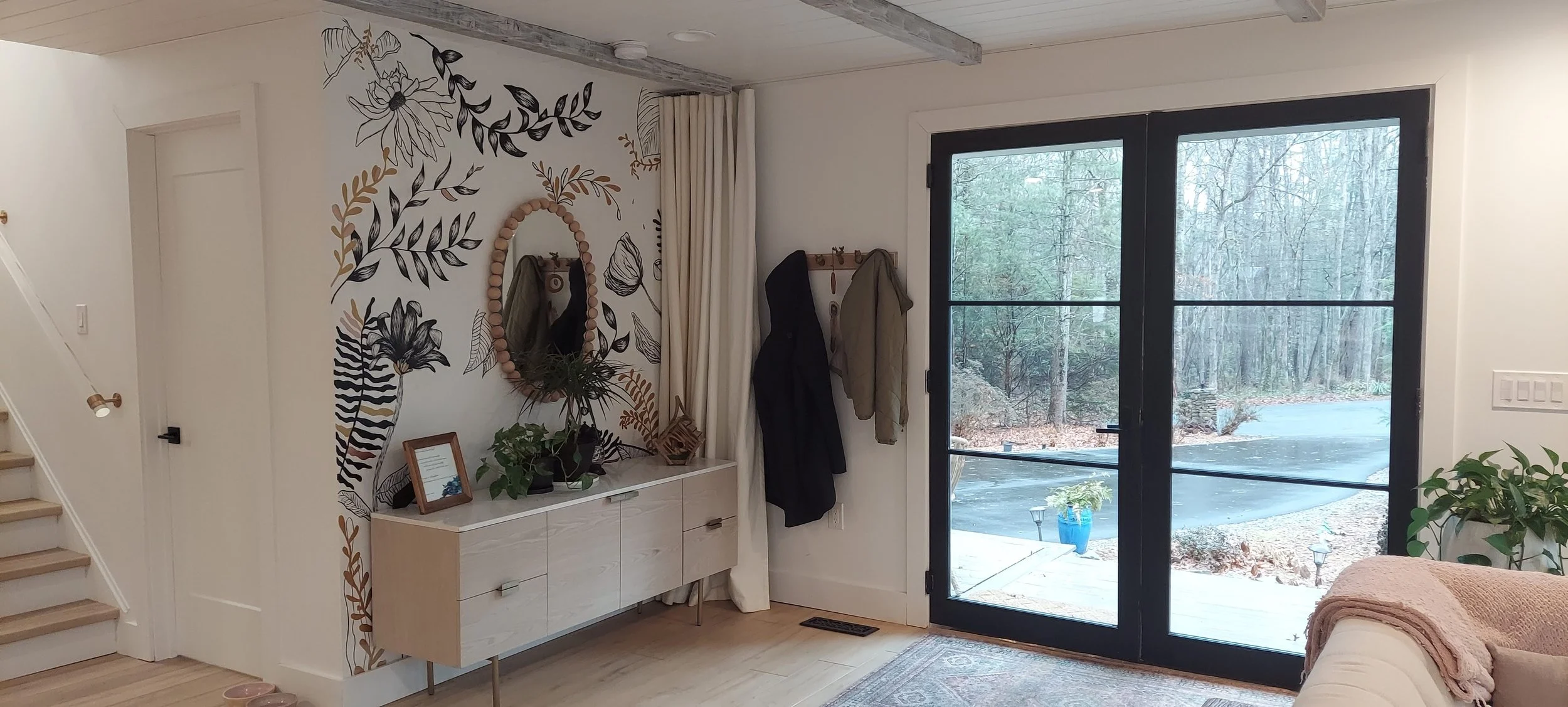 Interior of a modern home with a large glass door leading outside to a snowy yard, a wall with a black and gold floral mural, a white sideboard with plants and decorative items, and hanging coats near the door.