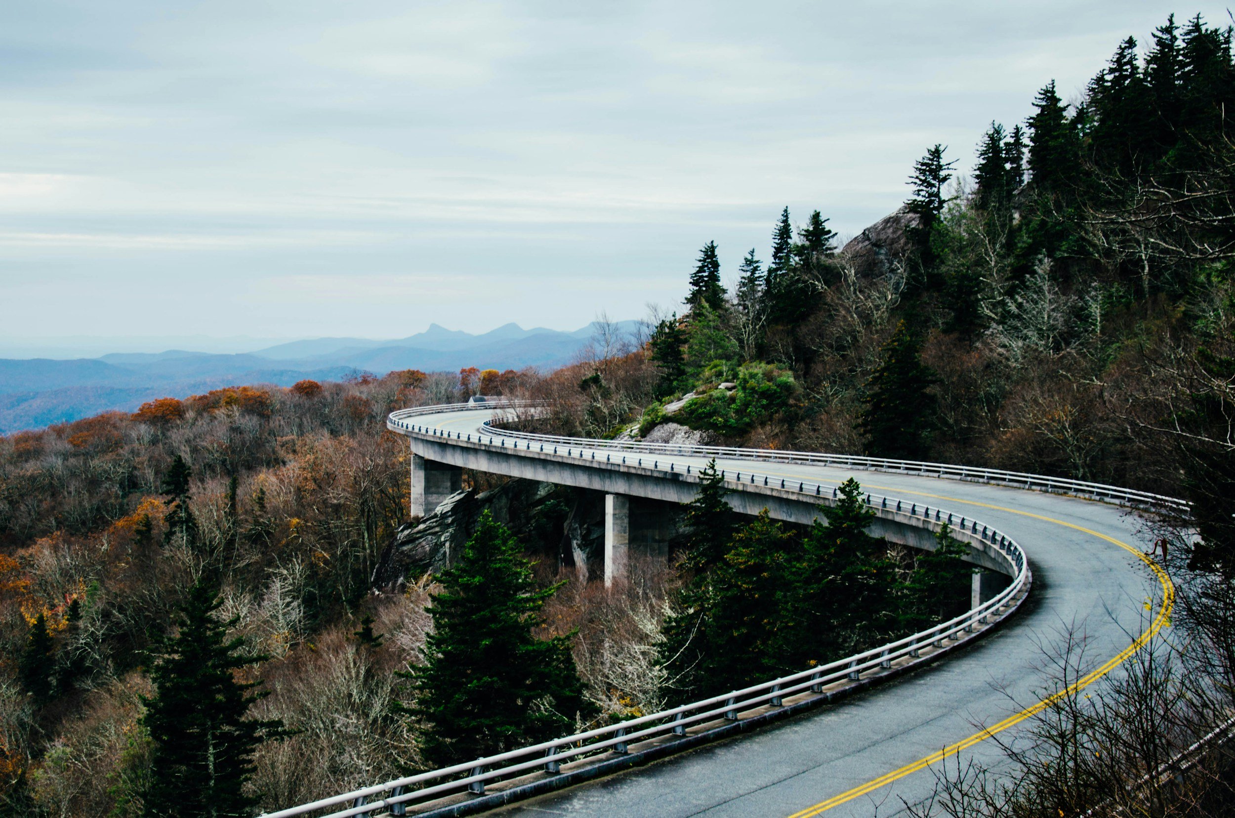 A winding mountain road with guardrails, surrounded by pine and deciduous trees, overlooking a landscape of distant mountains under a cloudy sky.