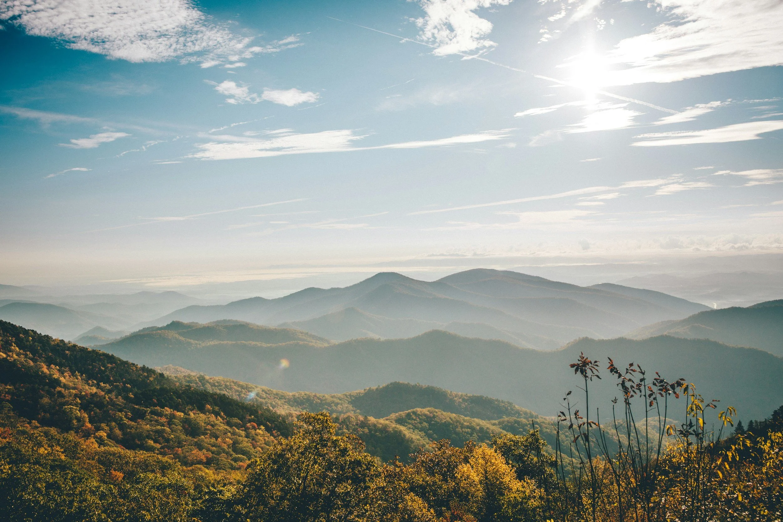 Scenic view of multiple layers of mountains with autumn-colored trees in the foreground, under a partly cloudy sky with the sun shining brightly.