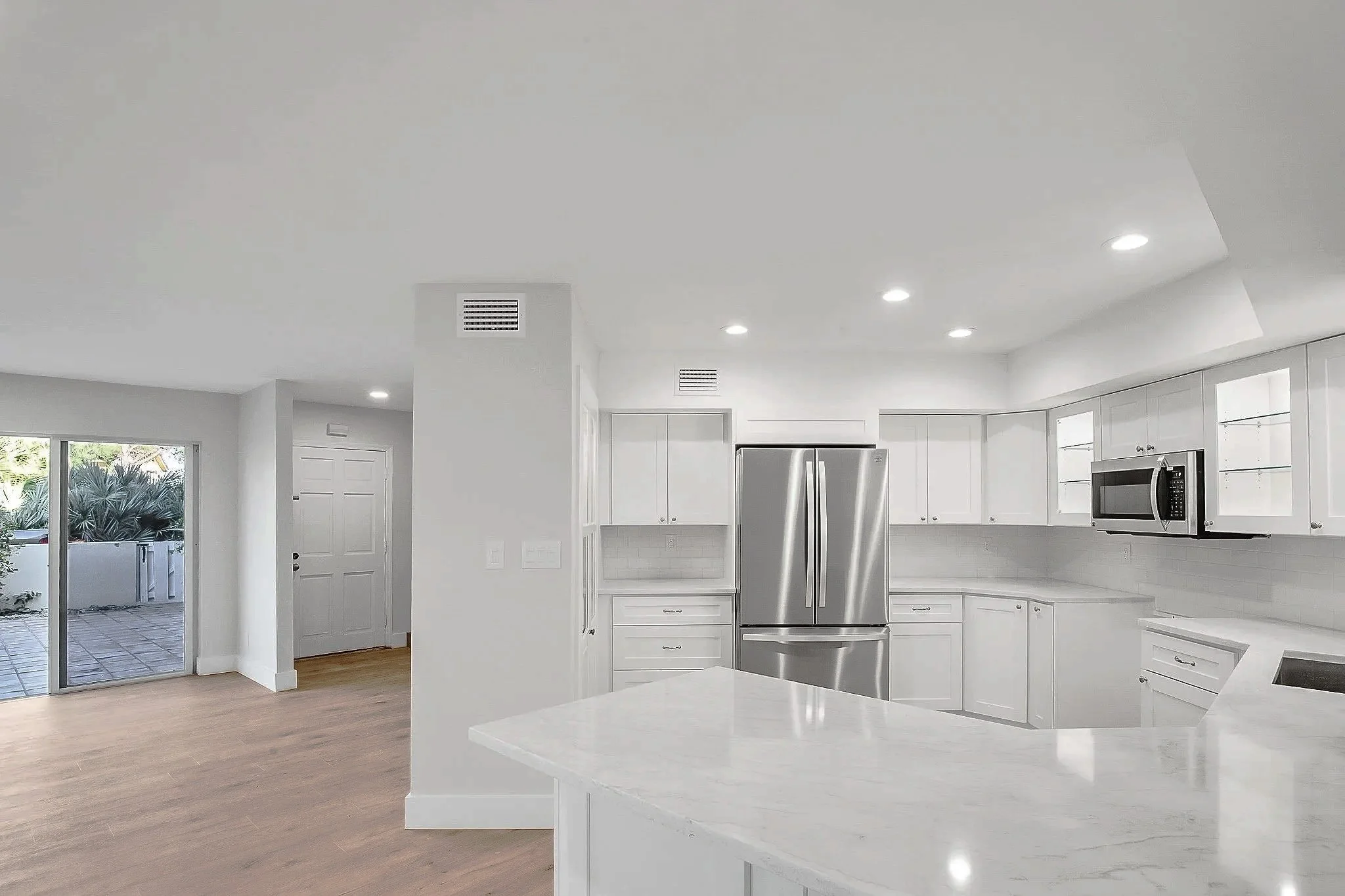 Modern kitchen with white cabinets, stainless steel refrigerator, microwave, recessed lighting, and a sliding glass door leading to an outdoor patio.