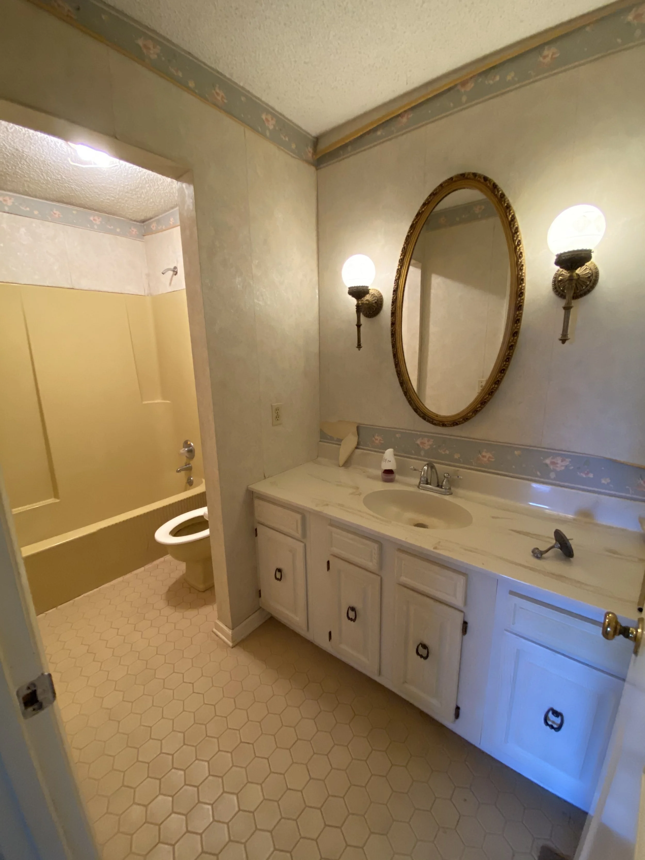 Bathroom with a vanity, oval mirror, two wall-mounted light fixtures, a yellow bathtub/shower combo, and a toilet in a small room with hexagonal tiled floor and floral wallpaper border.